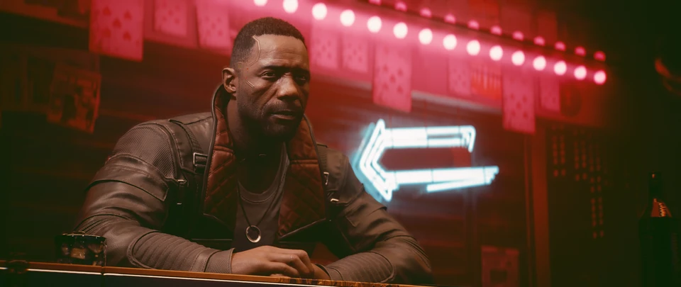 A man in a leather jacket sits at a table beside a neon arrow sign