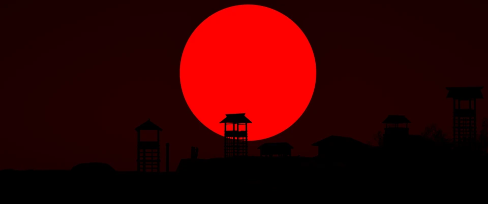 Large red sun over the silhouettes of a village and watchtowers against a black and red sky