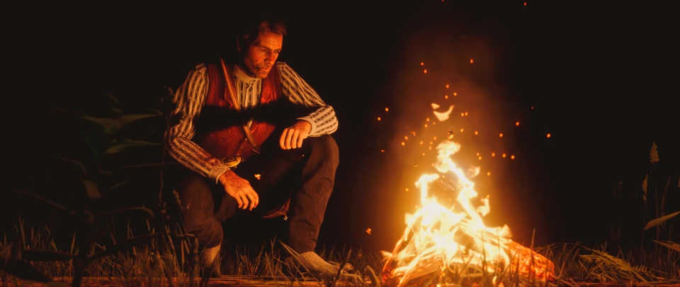 Man crouching beside a campfire at night as sparks rise